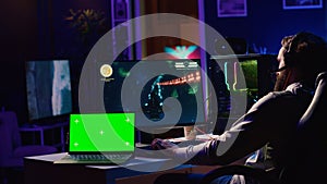 Green screen laptop next to man using gaming keyboard to play spaceship flying singleplayer game