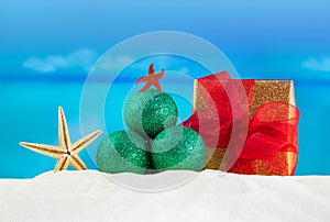 Green shiny Christmas tree made of glitter balls with starfish, wrapped gift box with red bow on sand of beach, sea behind
