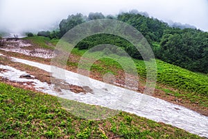 The green slopes of high mountains with melting snow are hidden in clouds and fog. Melting snow in high mountains in summer