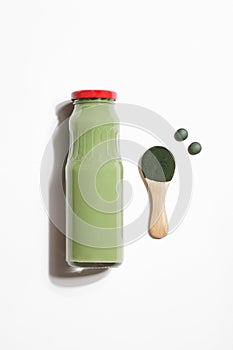 green smoothie in bottle and spirulina algae powder on wooden spoon and pills on white background