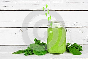 Green smoothie with kale and spinach in a mason jar with ingredients on a white wood background