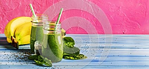 Green smoothies in glass bottles on cool pink blue background with yellow bananas. Panoramic banner with copy space