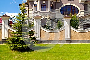 Green spruce grows on a green lawn in front of a wall of decorative stone in front of a rich house