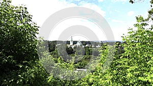 Green summer forest with city buildings behind. Clip. Blue cloudy sky and a small town behind trees.