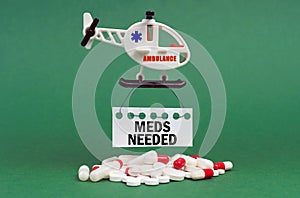 On a green surface, an ambulance helicopter, pills and a white sign with the inscription - Meds Needed