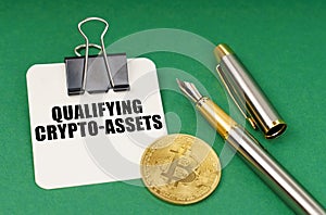 On a green surface, a bitcoin coin, a pen and a sheet of paper with the inscription - qualifying crypto-assets photo