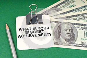 On a green surface, a pen, dollars and paper with the inscription - What Is Your Biggest Achievement