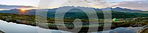 Green tent outdoors in the mountains by lake during sunset hours. Sunset/sunburst and clouds in the background, big panorama