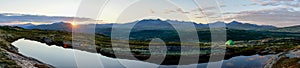 Green tent outdoors in the mountains by lake during sunset hours. Sunset/sunburst and clouds in the background, big panorama