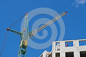 Green tower crane with yellow cabin and jib working on the construction site near building of white bricks