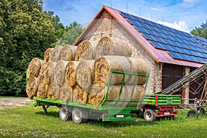 Green trailer filled with hay bales parked in the front of brick barn on a farm. Solar panels installed on the roof of the barn