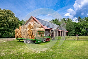 Green trailer filled with hay bales parked in the front of brick barn on a farm. Solar panels installed on the roof of the barn