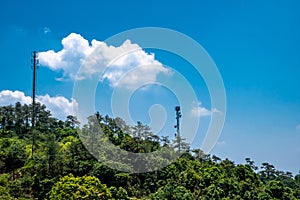 Green tree forest and telecommunication towers against clear blue sky with white fluffy clouds.