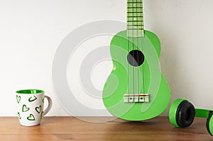 Green ukulele on a wooden table with a pair of headphones and a tea cup