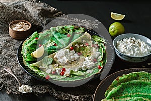 Green vegan crepes with spinach or pancakes with cottage cheese, pomegranate, nuts and spinach leaves on black plate over dark