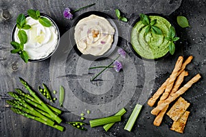 Green vegetables raw snack board with various dips. Yogurt sauce or labneh, hummus, herb hummus or pesto with crackers, grissini