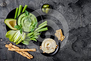 Green vegetables snack board with various dips. Hummus, herb hummus or pesto with crackers, grissini bread, fresh vegetables.