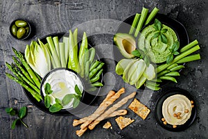 Green vegetables snack board with various dips. Yogurt sauce or labneh, hummus, herb hummus or pesto with crackers, grissini bread