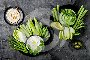 Green vegetables snack board with various dips. Yogurt sauce or labneh, hummus, herb hummus or pesto with fresh vegetables.