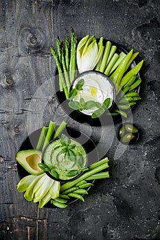 Green vegetables snack board with various dips. Yogurt sauce or labneh, hummus, herb hummus or pesto with fresh vegetables.