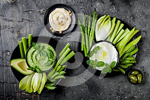 Green vegetables snack board with various dips. Yogurt sauce or labneh, hummus, herb hummus or pesto with fresh vegetables.