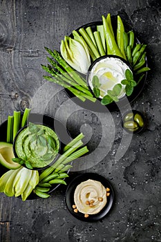 Green vegetables snack board with various dips. Yogurt sauce or labneh, hummus, herb hummus or pesto with fresh vegetables.