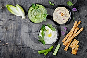 Green vegetables snack board with various dips. Yogurt sauce or labneh, hummus, herb hummus or pesto with fresh vegetables.