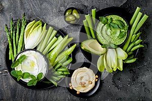 Green vegetables snack board with various dips. Yogurt sauce or labneh, hummus, herb hummus or pesto with fresh vegetables.