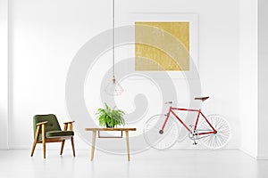 Green vintage armchair next to wooden coffee table with plant and red bike in living room interior with yellow painting on