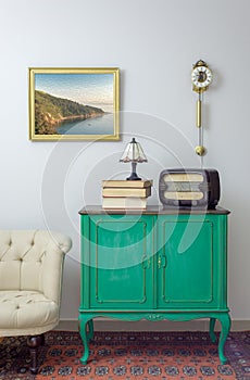 Green vintage sideboard with old radio and stack of old books, and table lamp on background of off white wall with hanged painting