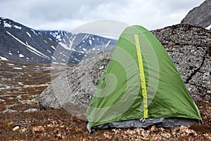 Green waterproof and windproof tent for hiking is in mountains at windy weather, standing behind big stone for protection from the