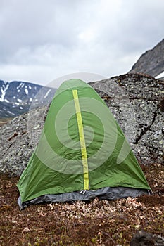 Green waterproof and windproof tent for hiking is in mountains at windy weather, standing behind big stone for protection from the