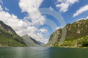 Green waters of the lake Lago d`Idro framed by alpine rocks with evergreen trees under a clear blue sky with white clouds