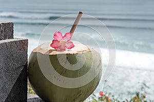 Green young fresh coconut close up with bamboo straw and pink tropical flower on the  cliff and blue ocean with big waves for