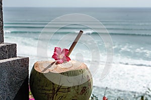 Green young fresh coconut close up with bamboo straw and pink tropical flower on the  cliff and blue ocean with big waves for