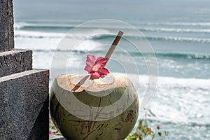 Green young fresh coconut close up with bamboo straw and pink tropical flower on the  cliff and blue ocean with big waves for