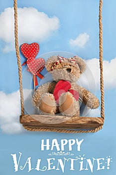 Greeting card with cute teddy bear swinging on a swing against blue sky and holding red heart for Valentine