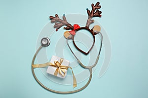 Greeting card for doctor with stethoscope, gift box and reindeer headband on light blue background, flat lay