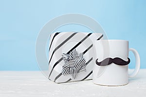 Greeting card fathers day holiday concept. White cup with mustache and gift box on blue pastel background