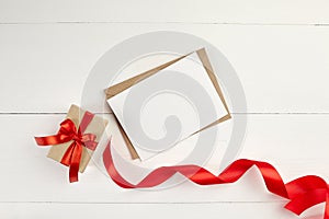 Greeting card mockup with paper envelope and red ribbon and gift box on white table