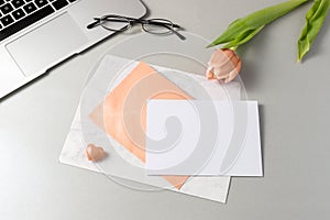 Greeting empty white card with envelope on grey table with peach fuzz color tulip flower and laptop