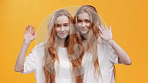 Greeting. Two young friendly redhead twin sisters waving hands hello to camera, bonding over orange studio background