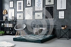 Grey armchair next to green mattress in bedroom interior with gallery of posters. Real photo