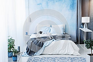 Grey blanket on bed in blue bedroom interior with plants and white lamp on table