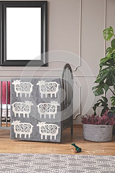 Grey blanket with sheep patter on the frame of grey wooden crib in elegant baby room with mockup poster on the wall