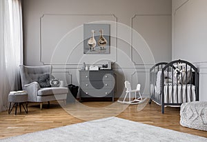 Grey chest of drawers in the middle of elegant grey baby room with comfortable armchair and wooden cradle
