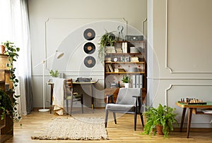 Grey comfortable armchair in vintage stylish interior with plants, book, and vinyls on the wall