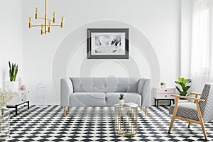 Grey couch and armchair on checkered floor in white flat interior with gold lamp and poster. Real photo