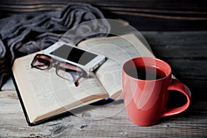 Grey cozy knitted scarf with cup of coffee or tea, phone, glasses and open book on a wooden table.