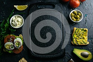 Grey cutting board with clear space. Around tomato, souce, olives, lemon, olive oil with spice and sandwiches
